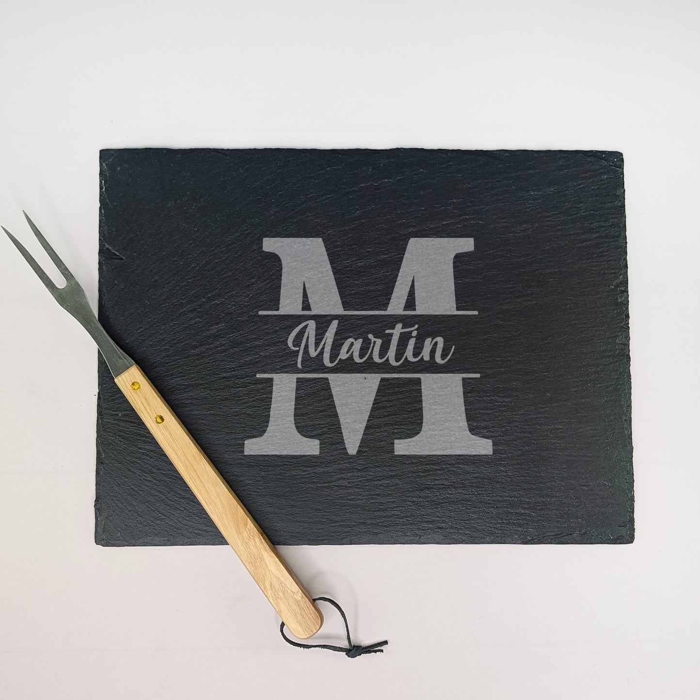 Schieferplatte mit Gravur „Martin“ und Initiale „M“, daneben Grillgabel mit Holzgriff – fotografiert von oben