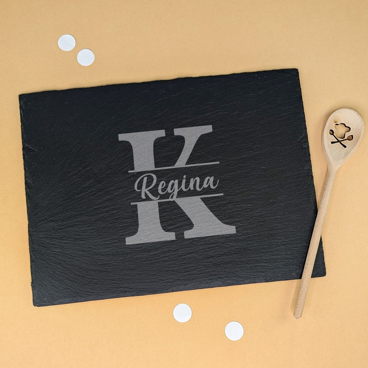 Schieferplatte mit Gravur „Regina“ und Initiale „K“, dekoriert mit Kochlöffel und Konfetti – auf beigem Hintergrund