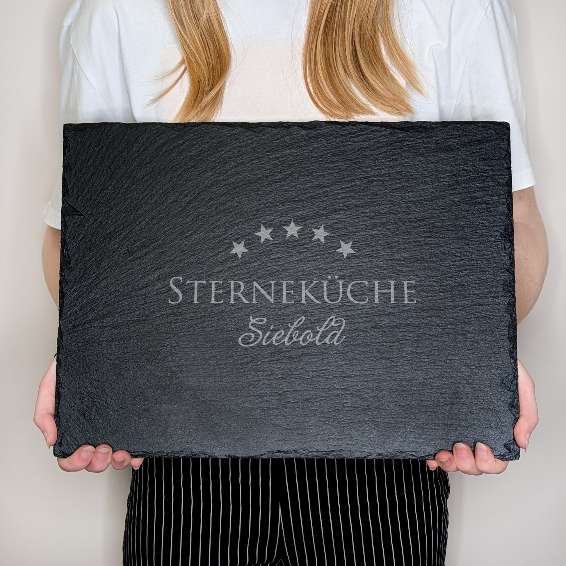 Schieferplatte mit Gravur „STERNEKÜCHE Siebold“, von einer Person in weißem Shirt und gestreifter Hose vor dem Körper gehalten – zur Größenansicht
