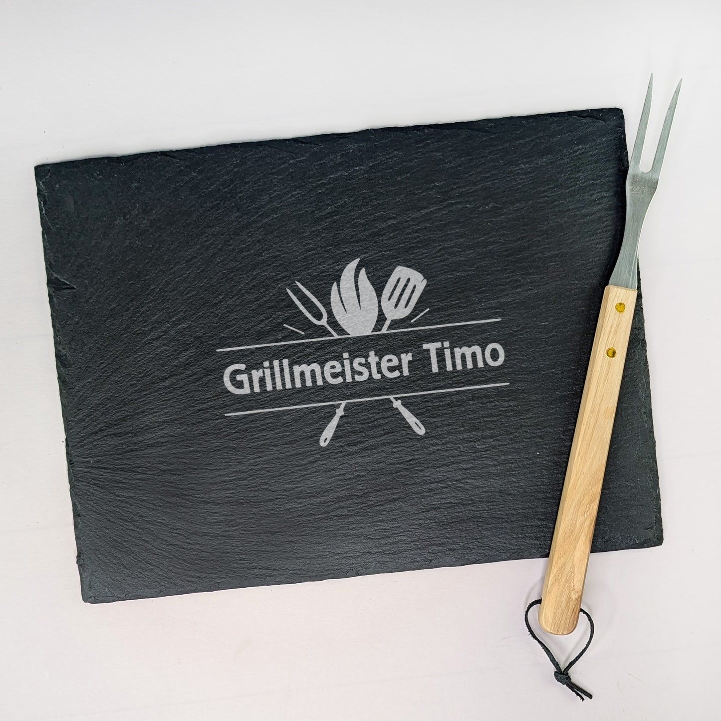 Schieferplatte mit Gravur „Grillmeister Timo“, daneben Grillgabel mit Holzgriff – fotografiert auf hellem Untergrund