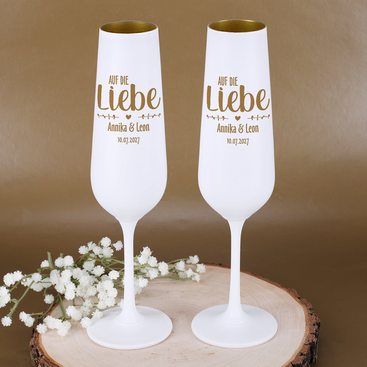 Zwei personalisierte Hochzeits-Sektgläser mit Gravur Auf die Liebe, Namen Annika & Leon und Datum 10.07.2027 auf einer Holzscheibe mit weißen Blumen