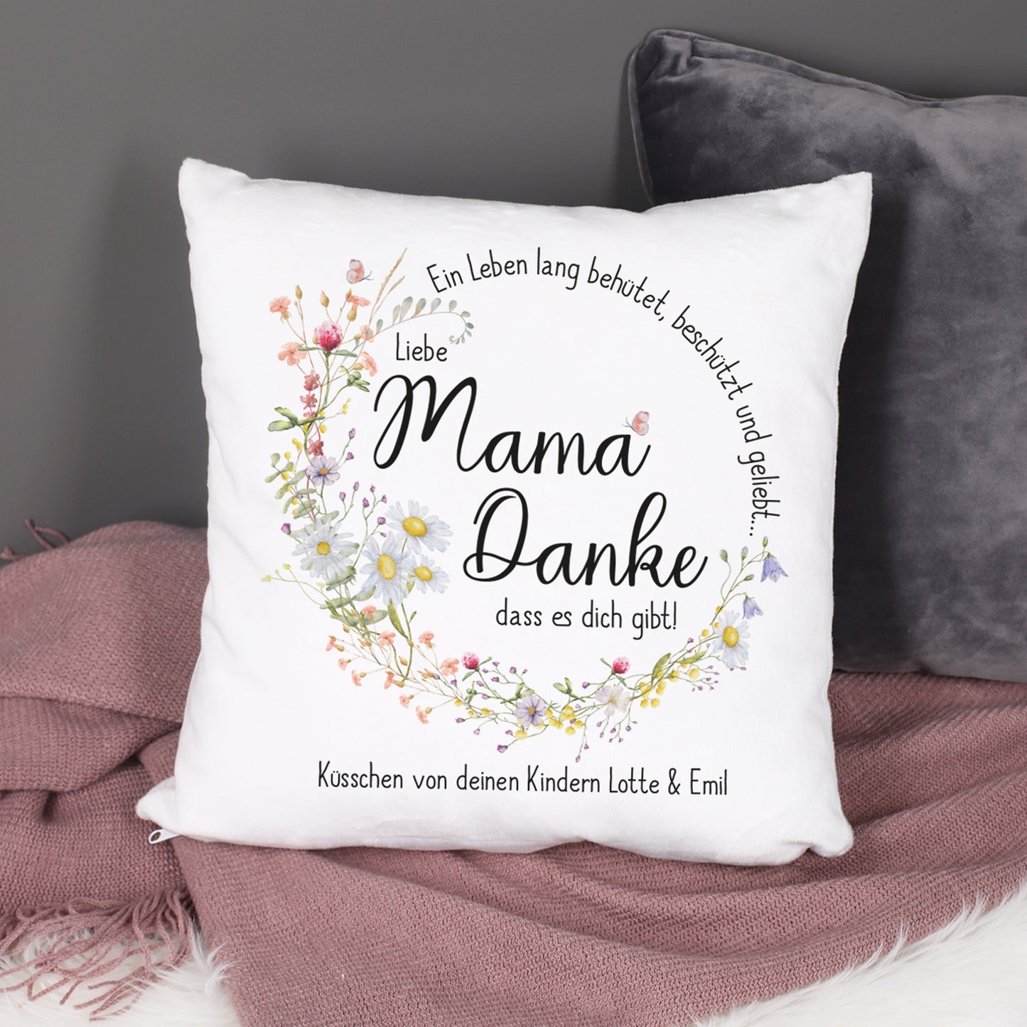Ein dekoratives Kissen für den Muttertag auf einem Sofa, umgeben von gemütlichen Kissen. Das Design zeigt einen floralen Wiesenblumenkranz mit dem Text „Mama Danke, dass es dich gibt!“.