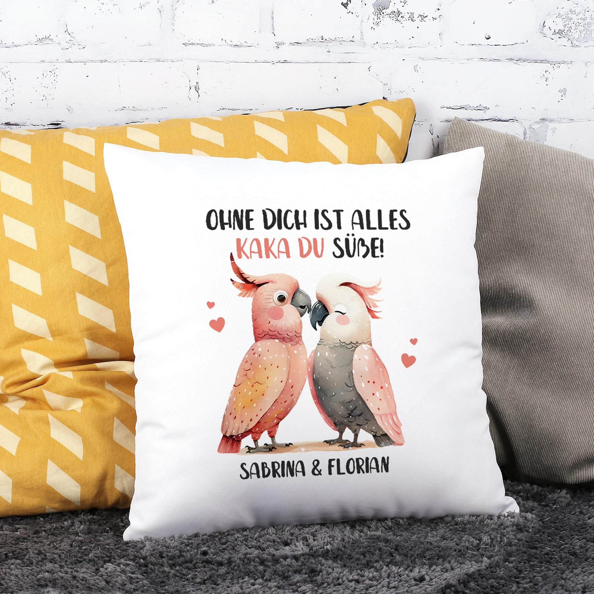 Personalisiertes Kakadu-Kissen mit romantischem Spruch und zwei Namen, dekoriert auf Sofa vor Backsteinwand