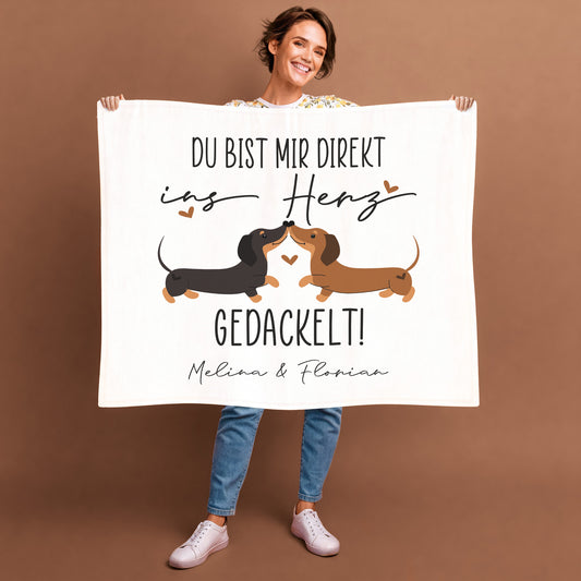 Personalisierte Kuscheldecke mit Dackel-Pärchen und Herzchen, Aufdruck „Du bist mir direkt ins Herz gedackelt!“ und Namen „Melina & Florian“, gehalten von Frau vor braunem Hintergrund