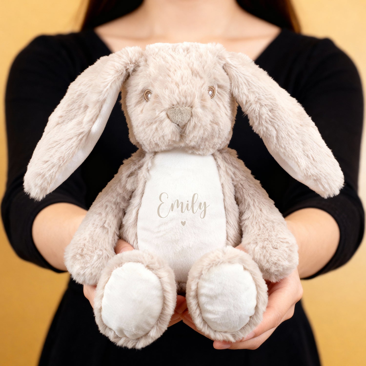Eine Person hält einen flauschigen beigefarbenen Hasen mit der Aufschrift „Emily“ und kleinem beigem Herzmotiv auf weißem Bauch. Das Kuscheltier hat lange Ohren und einen liebevollen Gesichtsausdruck.