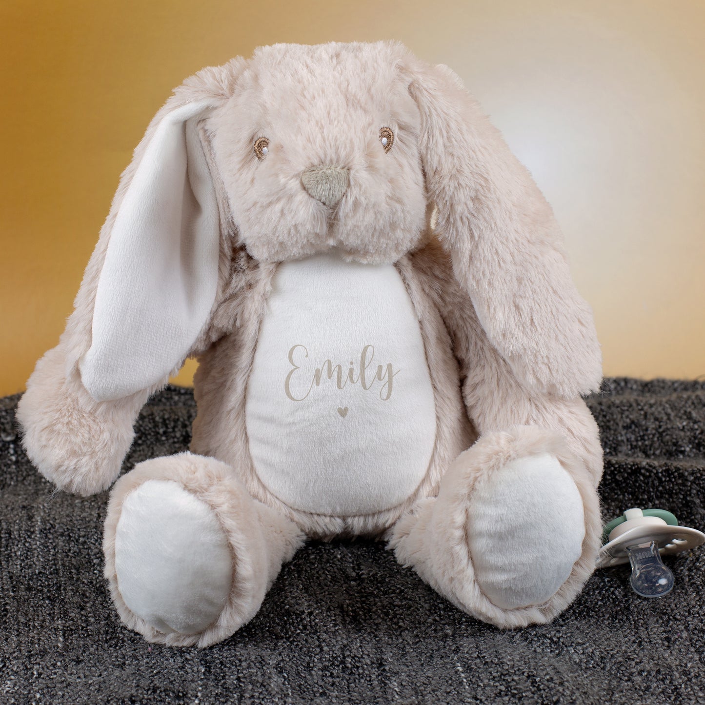 Beiger Kuschelhase mit Personalisierung „Emily“ in Beige sitzt auf grauer Decke. Daneben liegt ein Schnuller, der Hintergrund ist warm und goldfarben beleuchtet.