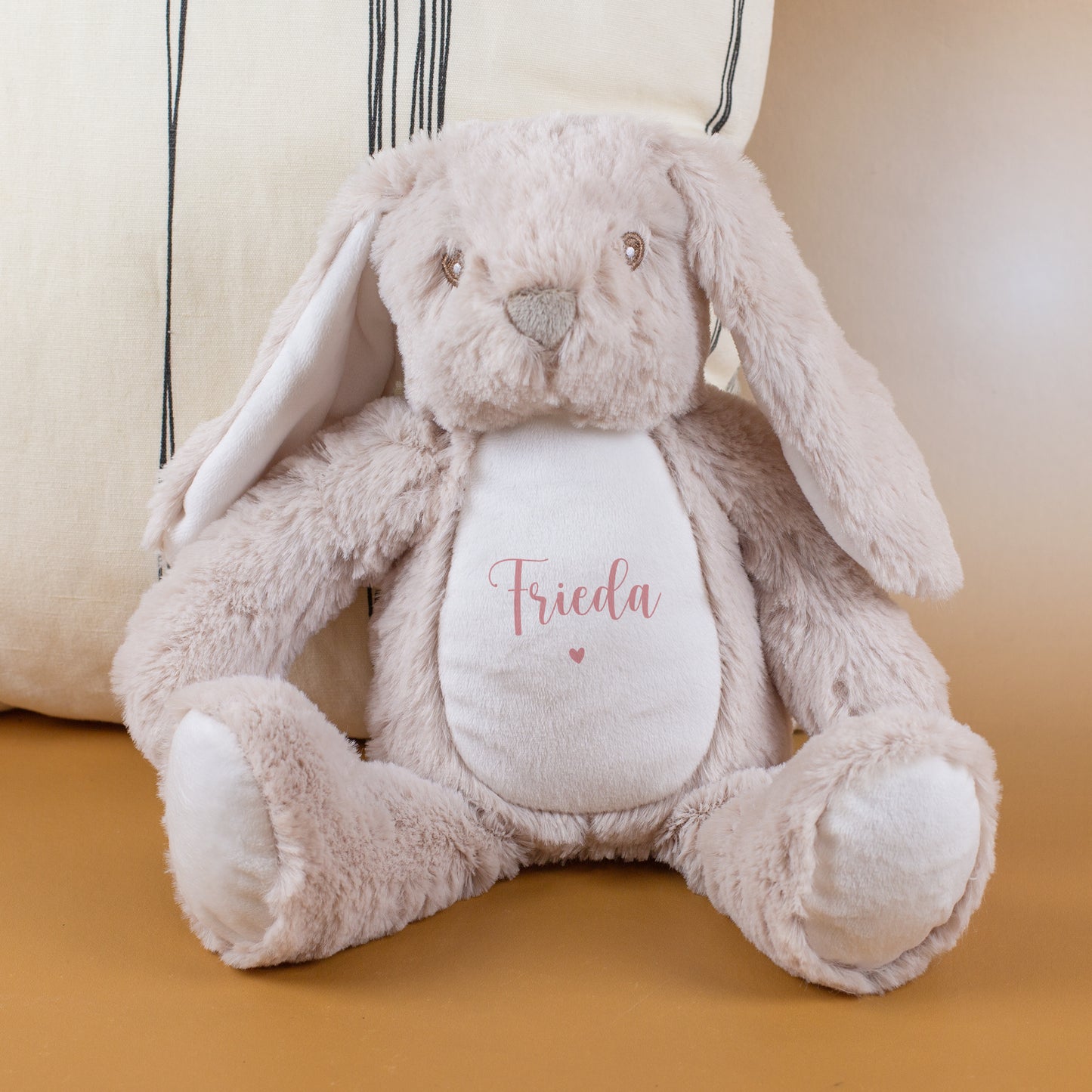Kuscheltier-Hase mit rosa Namensaufdruck „Frieda“ und Herzmotiv sitzt auf einer beigen Unterlage vor einem hellen Kissen. Das Plüschtier ist in Beige gehalten und hat weiche, lange Ohren.