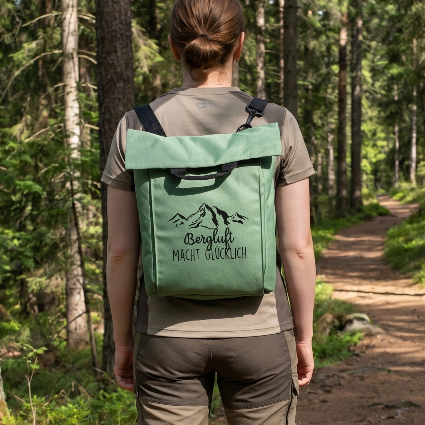 Jadegrüner Rucksack mit Bergmotiv und Wunschtext „Bergluft macht glücklich“ auf einem Waldweg