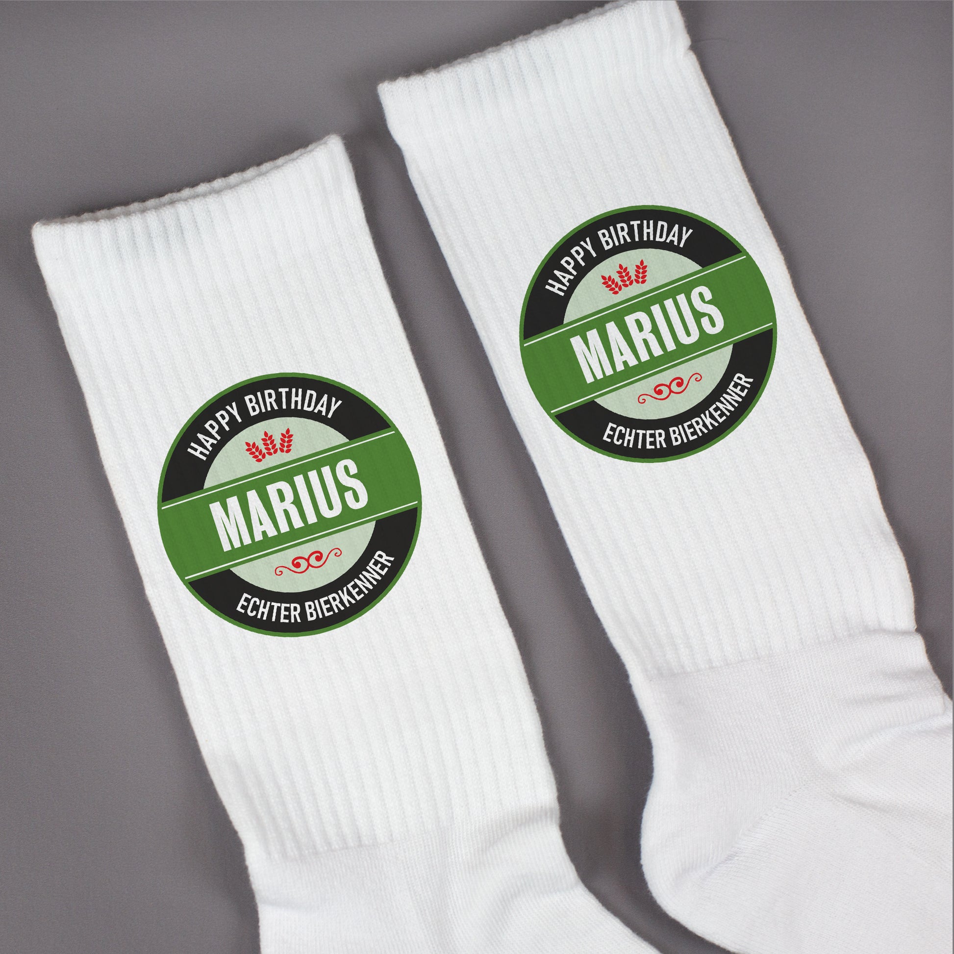Zwei weiße Socken mit personalisiertem Bierlogo „Marius – Echter Bierkenner“ – flachliegend auf grauer Fläche, Druckdetail.