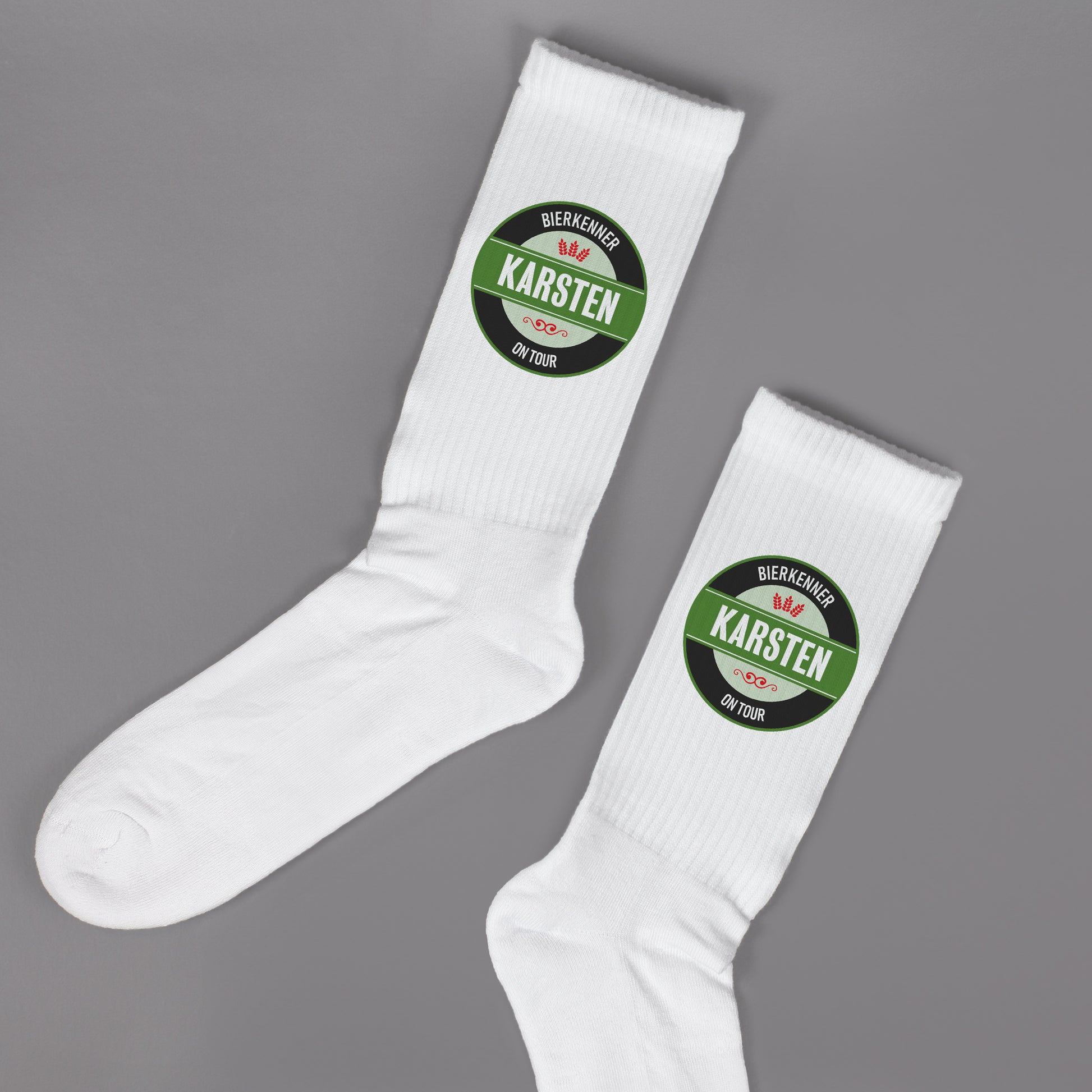Paar weiße Socken mit Bierlogo und Name „Karsten“ – Aufdruck im runden Etikettenstil, flachliegend auf grauem Untergrund.