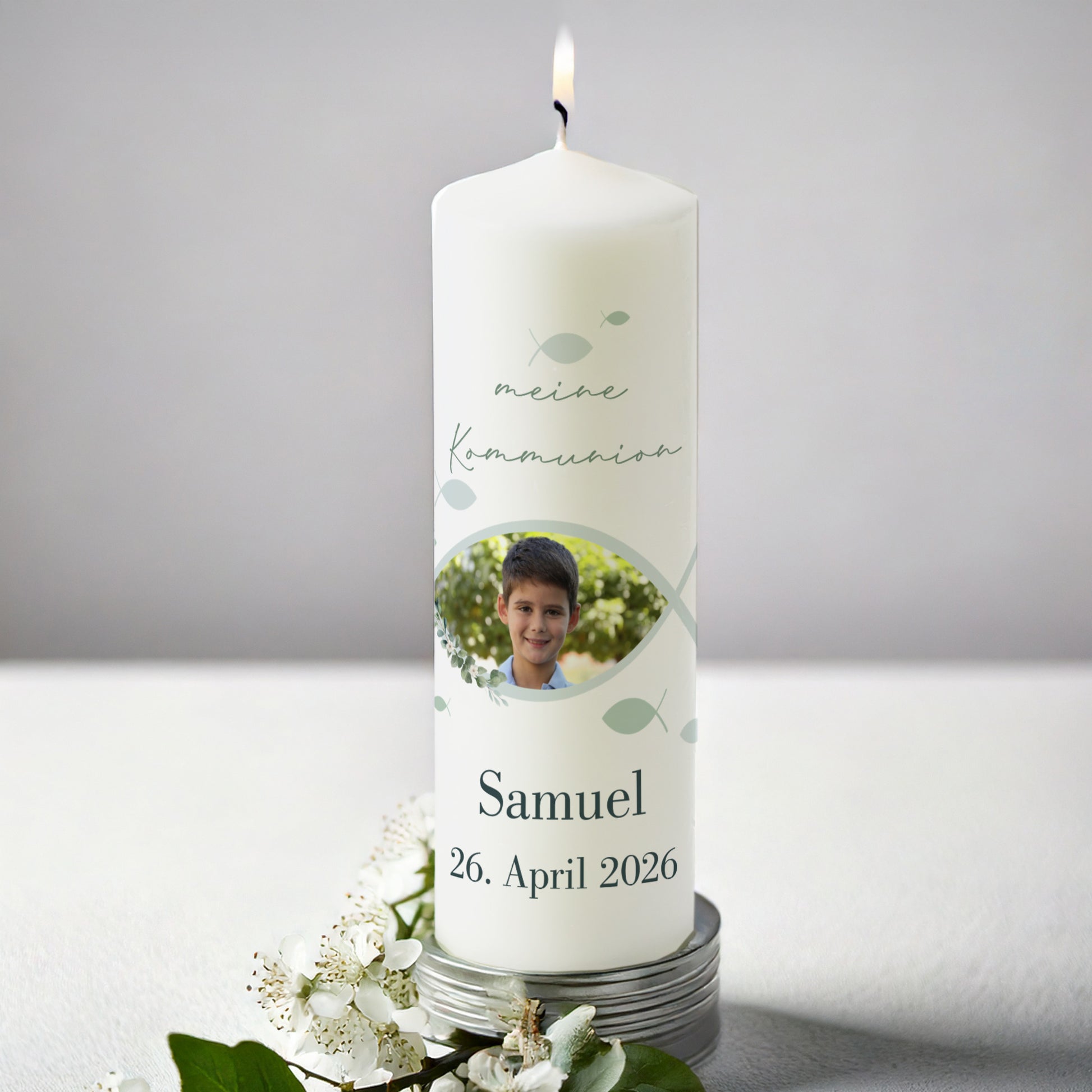 Weiße Kerze zur Kommunion mit Foto. Oben steht „meine Kommunion“, darunter das Foto eines Jungen, eingerahmt von einem stilisierten Fischsymbol. Darunter Name „Samuel“ und Datum „26. April 2026“. Die Kerze ist angezündet und von weißen Blüten umgeben.